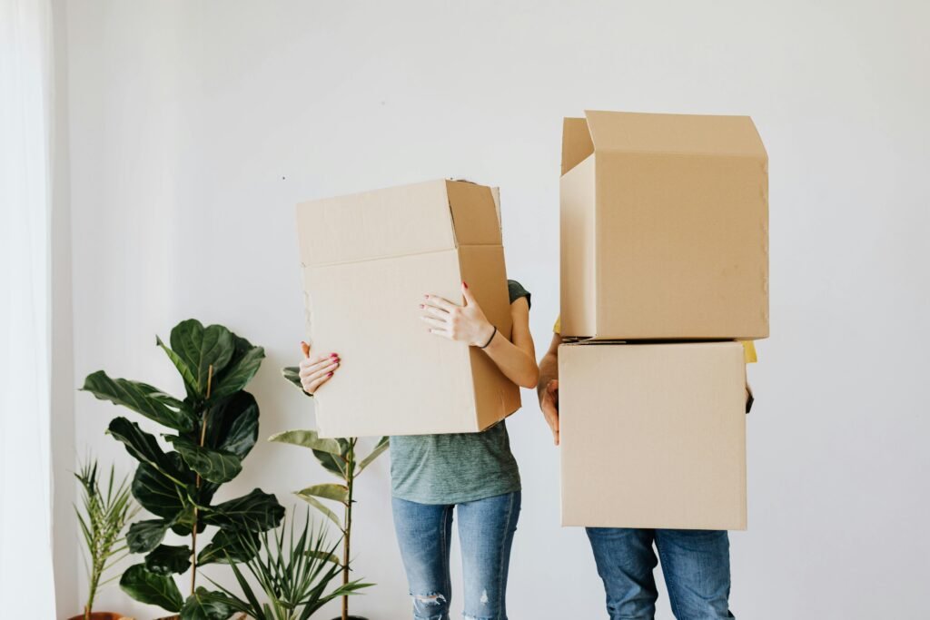 Unrecognizable couple wearing jeans standing carrying stacked carton boxes out of apartment during renovation on daytime